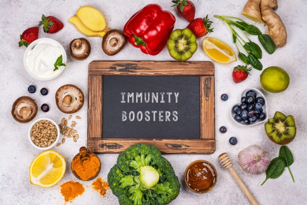 A small chalk board with the words "Immunity Boosters" written on it. The chalkboard is surrounded by various vegetables, fruits, yogurts, honey, and more.