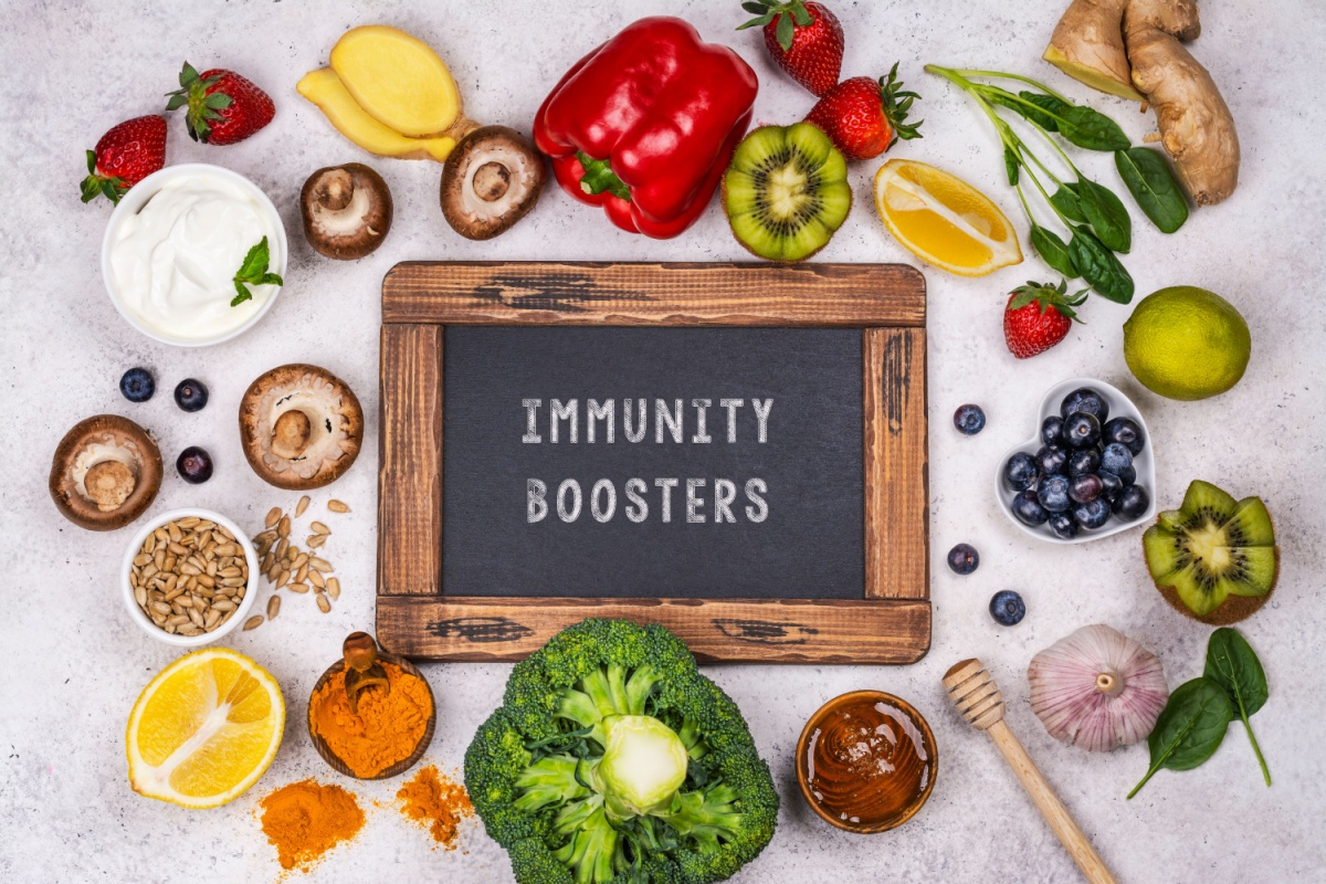 A small chalk board with the words "Immunity Boosters" written on it. The chalkboard is surrounded by various vegetables, fruits, yogurts, honey, and more.